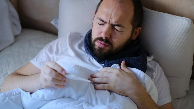 The man is sick in bed, checking the temperature. A person having a high fever watching a thermometer. The man is sick at home.