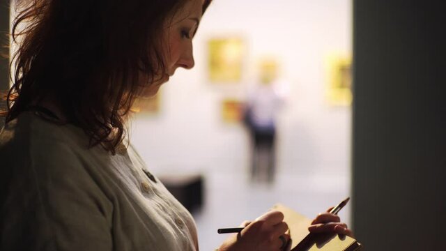 Silhouette female artist draws simple sketches in paper notebook with brown pages using pencil in dark hall of old art museum close-up shot