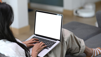 Close up view of young woman using mock up laptop computer with blank screen while sitting on sofa at home.