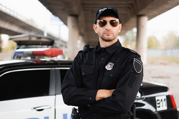 Police officer in sunglasses standing with crossed arms near auto on blurred background outdoors.