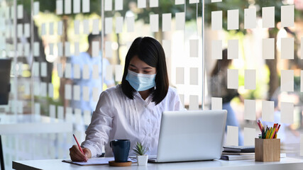 Female entrepreneur wearing face mask while working on a computer and writing notes in modern office.