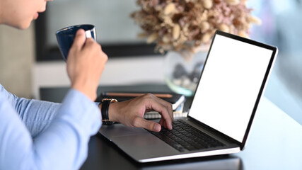 Cropped shot of young man using mock up laptop computer with blank screen and drinking coffee at office desk.