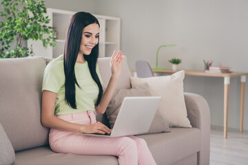 Photo of young pretty girl sit sofa hold netbook waving arm toothy smile wear green t-shirt pink pants indoors