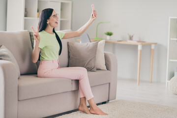 Obraz premium Photo of lovely girl sit sofa hold phone make selfie show v-sign open mouth barefoot wear specs green t-shirt pink pants indoors