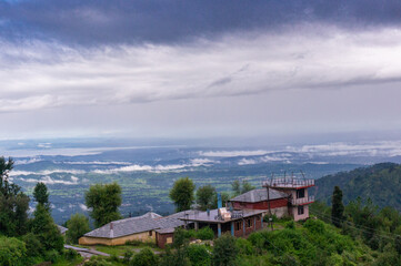 Fototapeta premium building perched on the edge of a mountain with trees all around looking over a beautiful vista landscape of a valley covered in clouds at dawn shot in Mcleodganj of dharamshala 