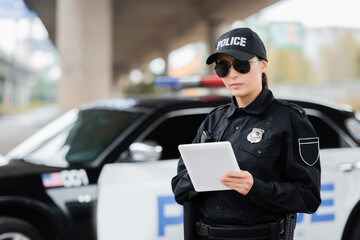 young policewoman with tablet looking at camera with blurred patrol car on background outdoors.