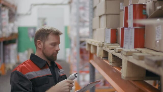 Medium close-up of caucasian construction contractor in blue overall scanning barcodes on boxes with goods on pallet racking at warehouse with handheld scanner