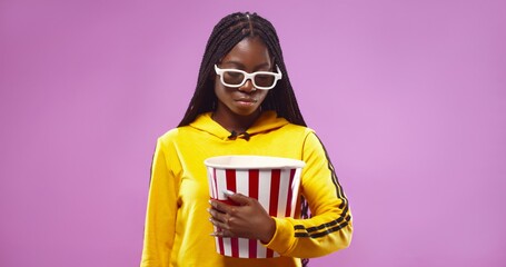 Portrait of young African American woman in 3d glasses holding bucket with popcorn isolated over pink background. Cinema concept