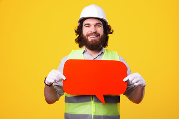 Cheerful bearded architect smiling and holding empty speech bubble over yellow background.