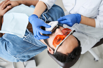 Dentist whitening teeth of bearded man sitting in dental chair