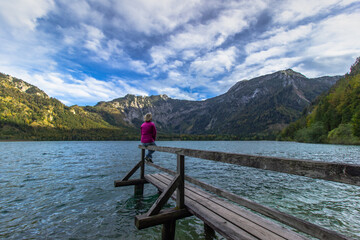 Fototapeta na wymiar Young girl having a rest by mountain lake, Austria. Female traveler enjoying view of Alps. Wanderlust freedom travel concept. Summer vacation adventure scene. Sitting relaxing woman on her holiday