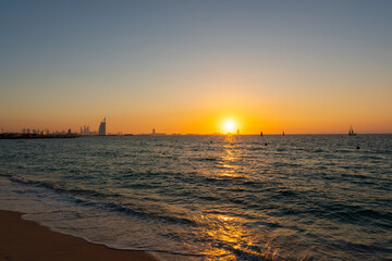 Amazing orange sunset at the beach in Dubai. Beautiful orange colors coming from sun and reflect over Gulf sea waves and shore of UAE. Silhouette of the iconic hotels visible at the horizon line. 