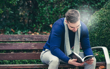 young man sitting on a park bench reading a book