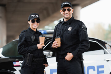 Smiling police officers in sunglasses holding takeaway coffee near car on blurred background...