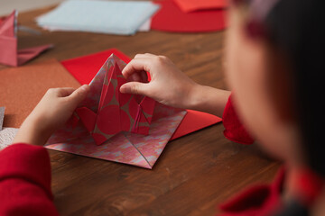 Obraz na płótnie Canvas Unrecognizable little Asian girl sitting at wooden table making origami crane using colored paper, ocer-the-shoulder shot
