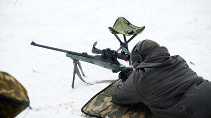 A sniper in position looks through a telescopic sight at a sporting event. Near a chair