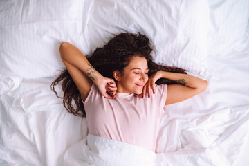 Top view of young woman waking up in early morning lying on bed with whitesnow linen and stretch...