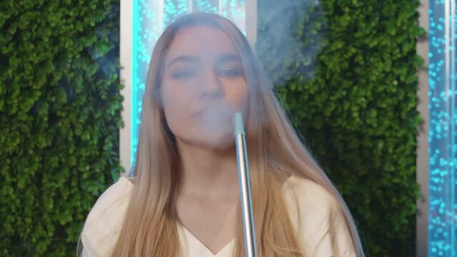 Young blonde woman smokes hookah and looks at the camera