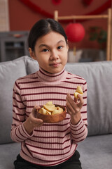 Medium portrait of young Asian girl sitting on sofa in living room choosing fortune cookie looking...