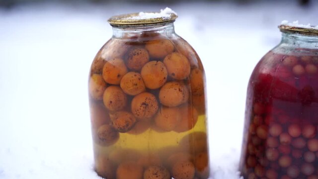 Close up of canned juice with fruits and berries in snow on street. Sealed jars of compote in garden in wintertime.