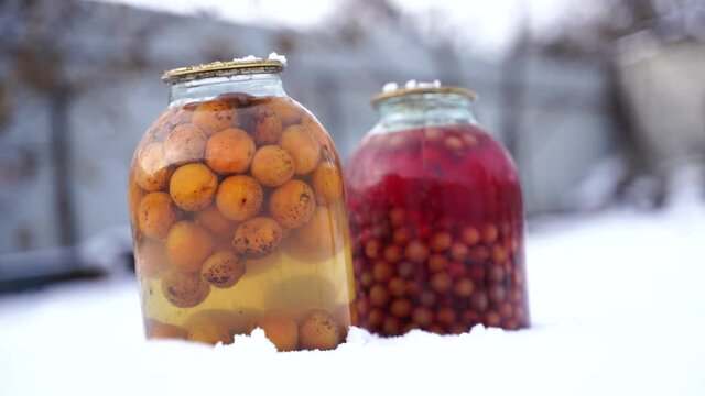 Close up of canned juice with fruits and berries in snow on street. Sealed jars of compote in garden in wintertime.