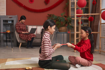 Two little Chinese girls sitting on floor in front of each other holding red envelope for Lunar New Year celebration, their mother using laptop on background