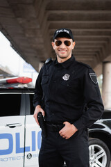 Smiling police officer in sunglasses holding gun in holster near car on blurred background.