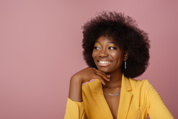 Yong beautiful happy smiling African American woman, model wearing elegant jewelry, yellow blazer, posing in studio, on pink background. Girl looking aside. Copy, empty space for text
