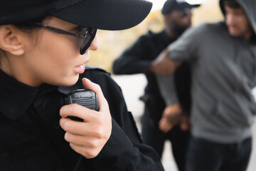 close up view of policewoman talking on radio set during african american colleague arresting offender on blurred background.