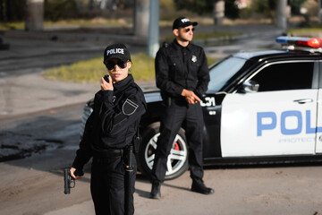 Fototapeta na wymiar Policewoman with gun using walkie talkie near colleague and police car on blurred background on urban street.