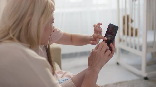 Over-the-shoulder close-up of pregnant woman showing ultrasound image of baby on smartphone to mother talking about future baby together sitting at home