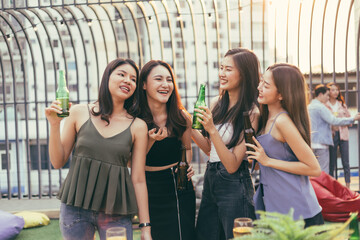 Group of happy Asian girl friends celebrating party with beer bottle toasting drinks at rooftop cafe in sunset together, female gang chatting, laughing on smile face, night lifestyle of young people