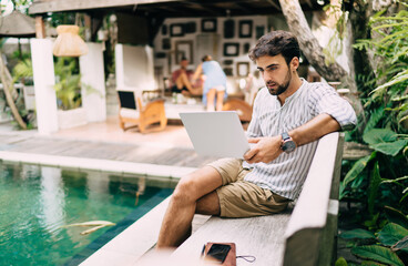Concentrated man working on laptop on poolside in tropical resort