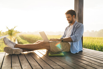 Cheerful remote employee typing on laptop on porch in sunlight