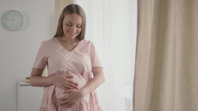 Medium slowmo portrait of smiling young caucasian pregnant woman stroking belly looking at camera in bright living room