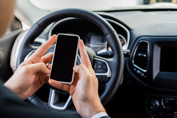 Cropped view of man holding smartphone with blank screen near steering wheel in car on blurred background.