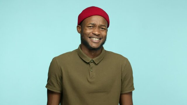 Slow motion of african american guy in beanie looks with cringe and dislike at camera, rejecting offer and smiling, showing teeth braces, standing over turquoise background