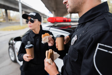 Policeman holding burger and coffee to go near colleague and car on blurred background on urban street.