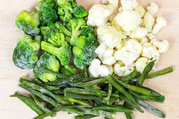 Top view of different frozen vegetables (broccoli, green beans, cauliflower) on light wooden background.
