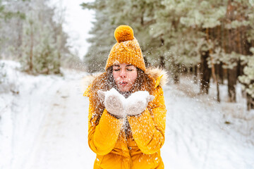Beautiful young woman in yellow jacket and hat throws snow in snowy winter forest
