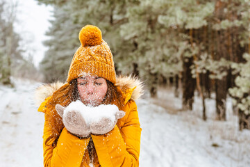 Outdoor close-up portrait of a young stylish smiling smiling girl wearing a yellow jacket and a knitted hat, walking in the winter forest and playing with the snow outdoors. Christmas holidays