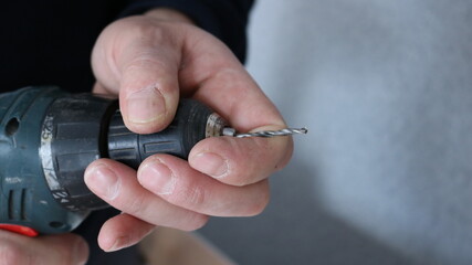 close-up carpenter hands holding green drill body with red button, thin drill setting by male working hands on gray blank wall background with place for text
