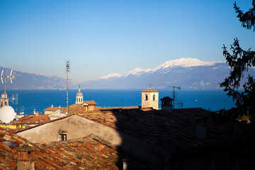 Fototapeta na wymiar Aerial view of the snow-capped Italian alps, Garda lake and small european buildings with orange tiled roofs, towers and chimneys. Panorama of Desenzano del Garda, Lombardy, Italy.
