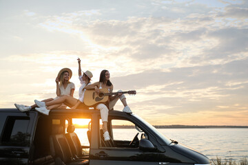 Happy friends with guitar sitting on car roof outdoors at sunset. Summer trip