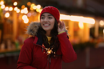 Happy young woman with sparkler at winter fair in evening. Christmas celebration