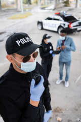 Policeman in latex gloves and medical mask using walkie talkie while colleague talking with african american victim on blurred background on urban street.