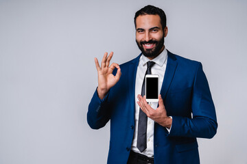 Smiling confident Arab businessman showing okay sign and smart phone isolated over grey background