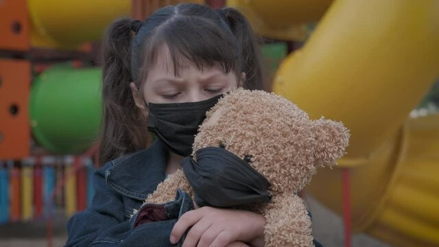 Sits on playground in mask. An alone child with teddy bear both in medical mask stay on the playground during quarantine.