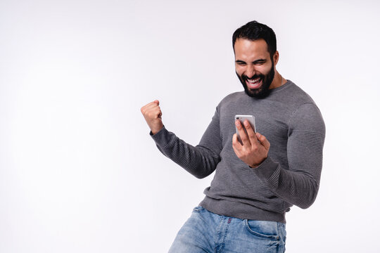 Happy excited young Arabian man in casual attire using mobile phone isolated over white background