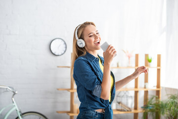  blonde woman in headphones singing while holding smartphone at home on blurred background